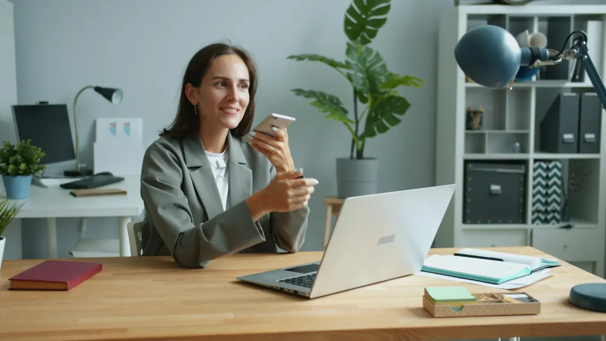 A woman speaking into her phone at a desk beside a laptop